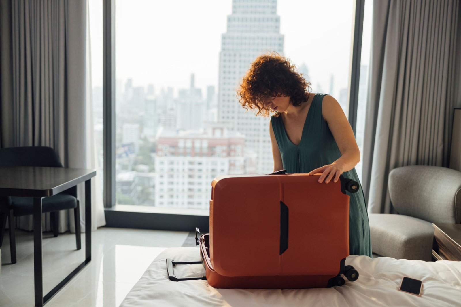 A woman unpacking a large suitcase in a high-rise apartment after moving abroad alone, with a city skyline outside