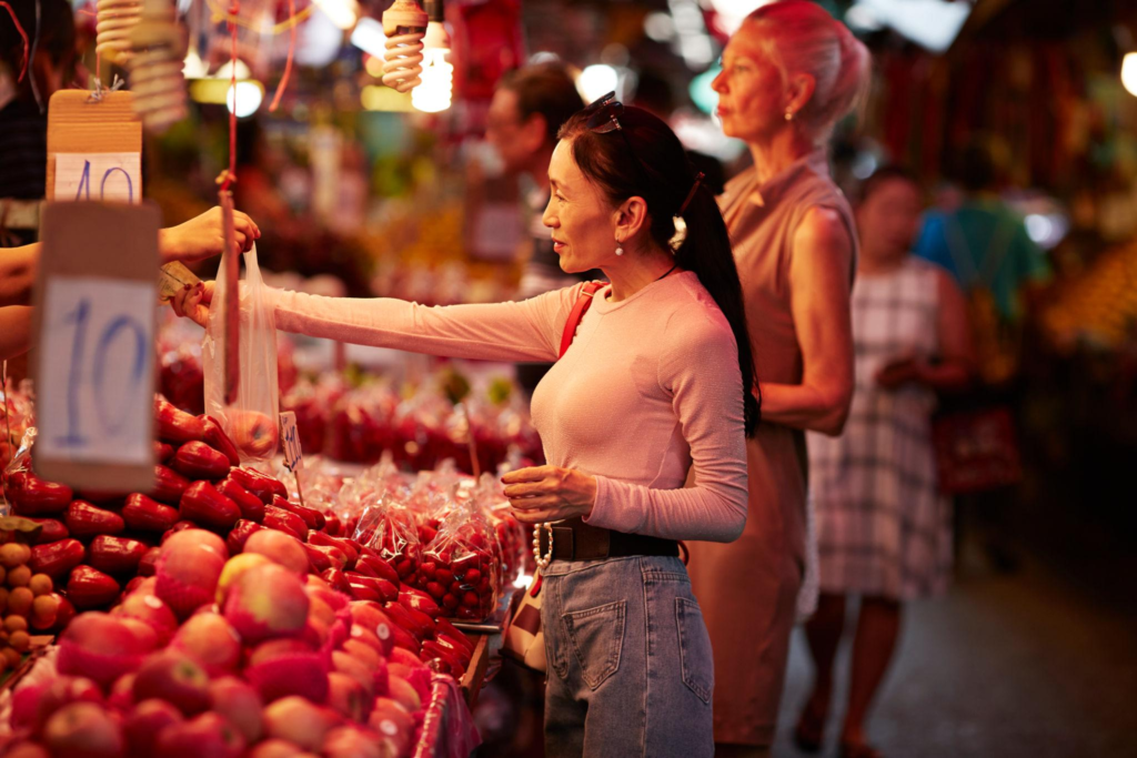 A woman paying cash and receiving fruit at a busy local market, representing everyday access to money while living abroad